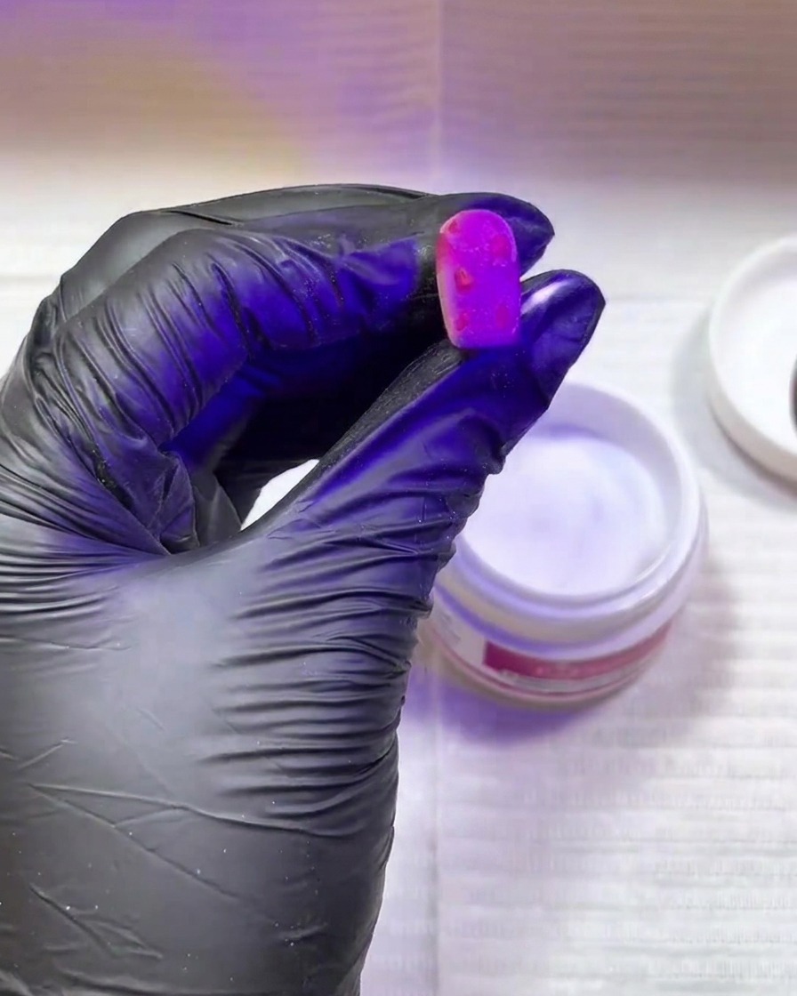 Mano con guante negro curando una uña rosa con manchas texturizadas bajo luz UV.