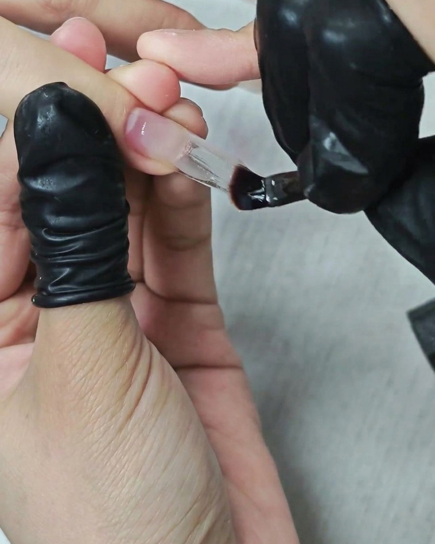 Una manicurista usando un pincel plano para aplicar y dar forma a gel constructor transparente grueso sobre una extensión de uña cuadrada.