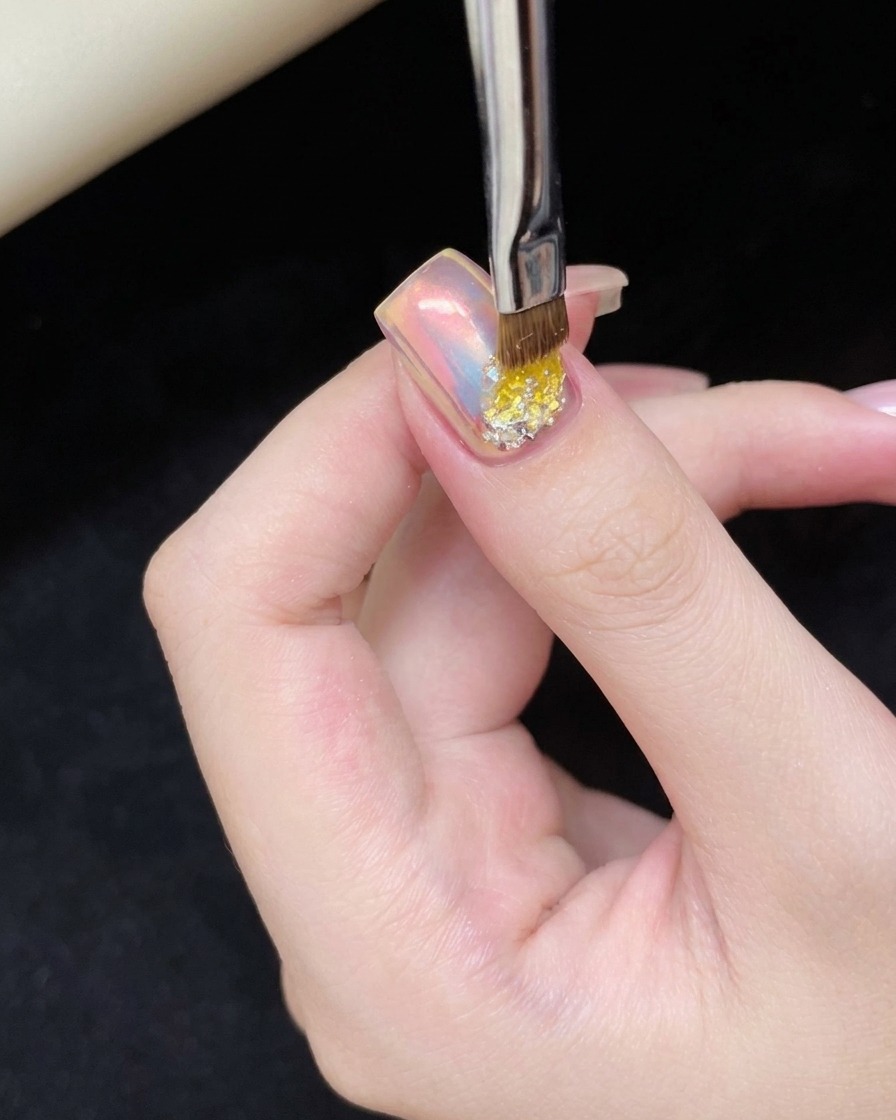 Un pequeño pincel de uñas aplicando un toque de gel adhesivo transparente en la parte inferior de una uña ojo de gato rosa.