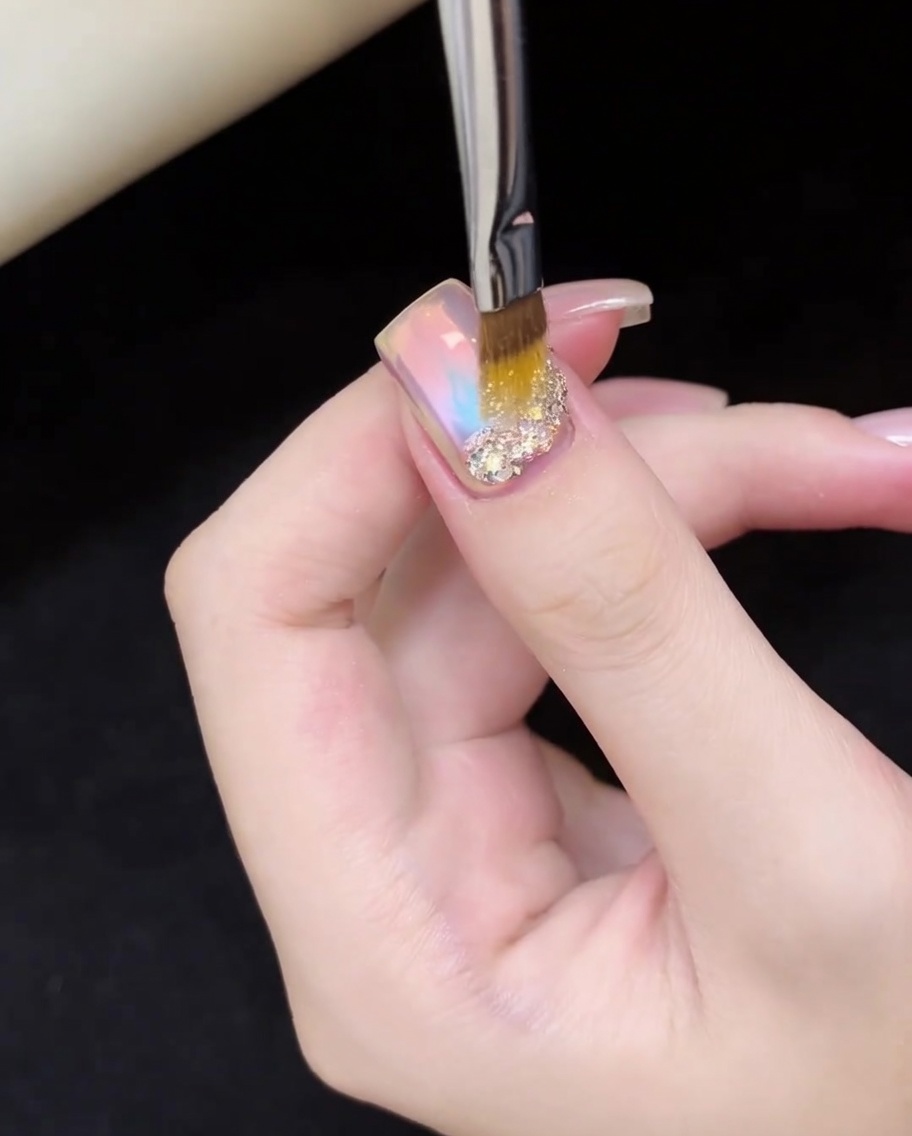 Un pincel de uñas colocando con precisión escamas de glitter dorado y plateado iridiscente en la base de la uña.