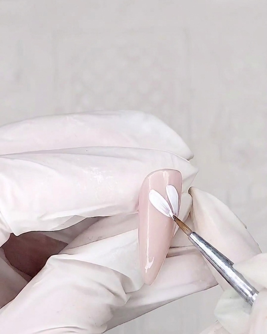Un pincel de detalle pintando pétalos blancos sobre una uña con base rosa.