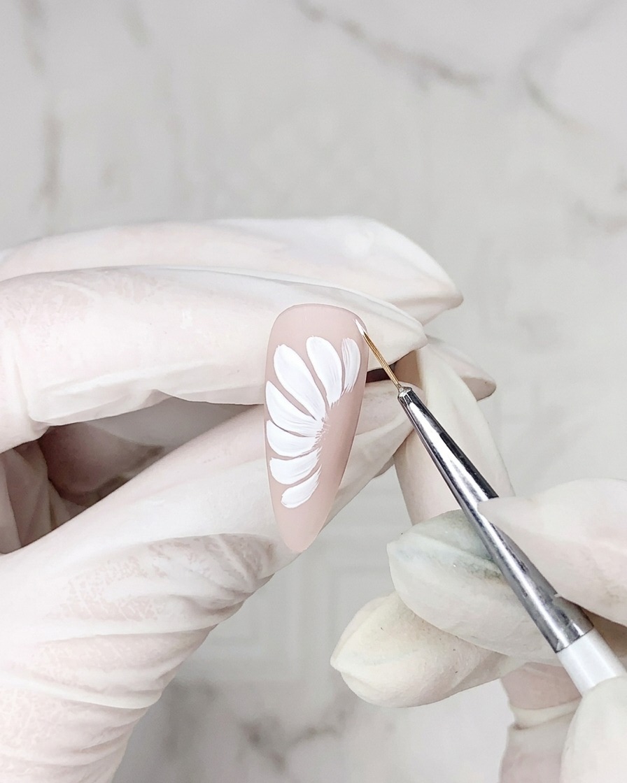 Primer plano de un pincel de detalle aplicando una línea fina de gel adhesivo alrededor del borde de un pétalo de margarita blanco sobre una base de uña nude.