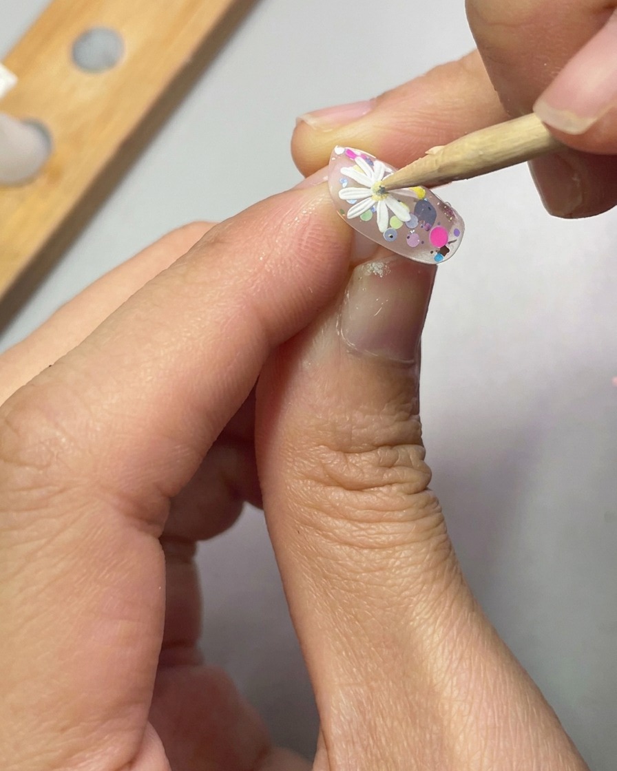 Una herramienta de punteado colocando una pequeña gota de gel amarillo en el centro de una margarita blanca 3D sobre una uña decorada.