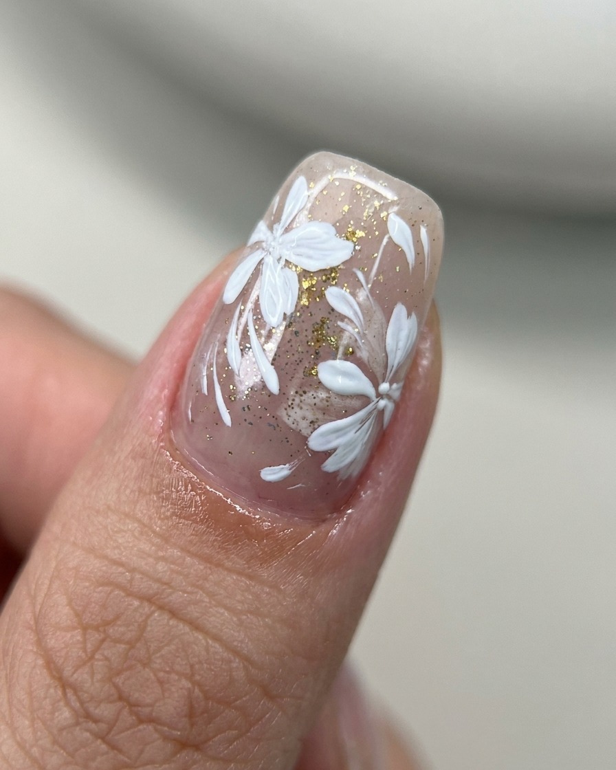 Un pincel aplicando top coat transparente sobre una uña decorada con arte floral y acentos dorados.