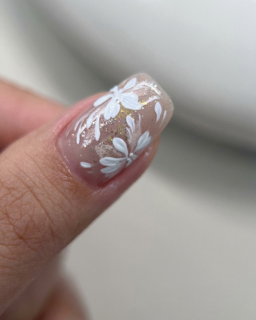 Primer plano de un pincel liner añadiendo pequeños pétalos blancos a un diseño floral existente en una uña de gel nude.