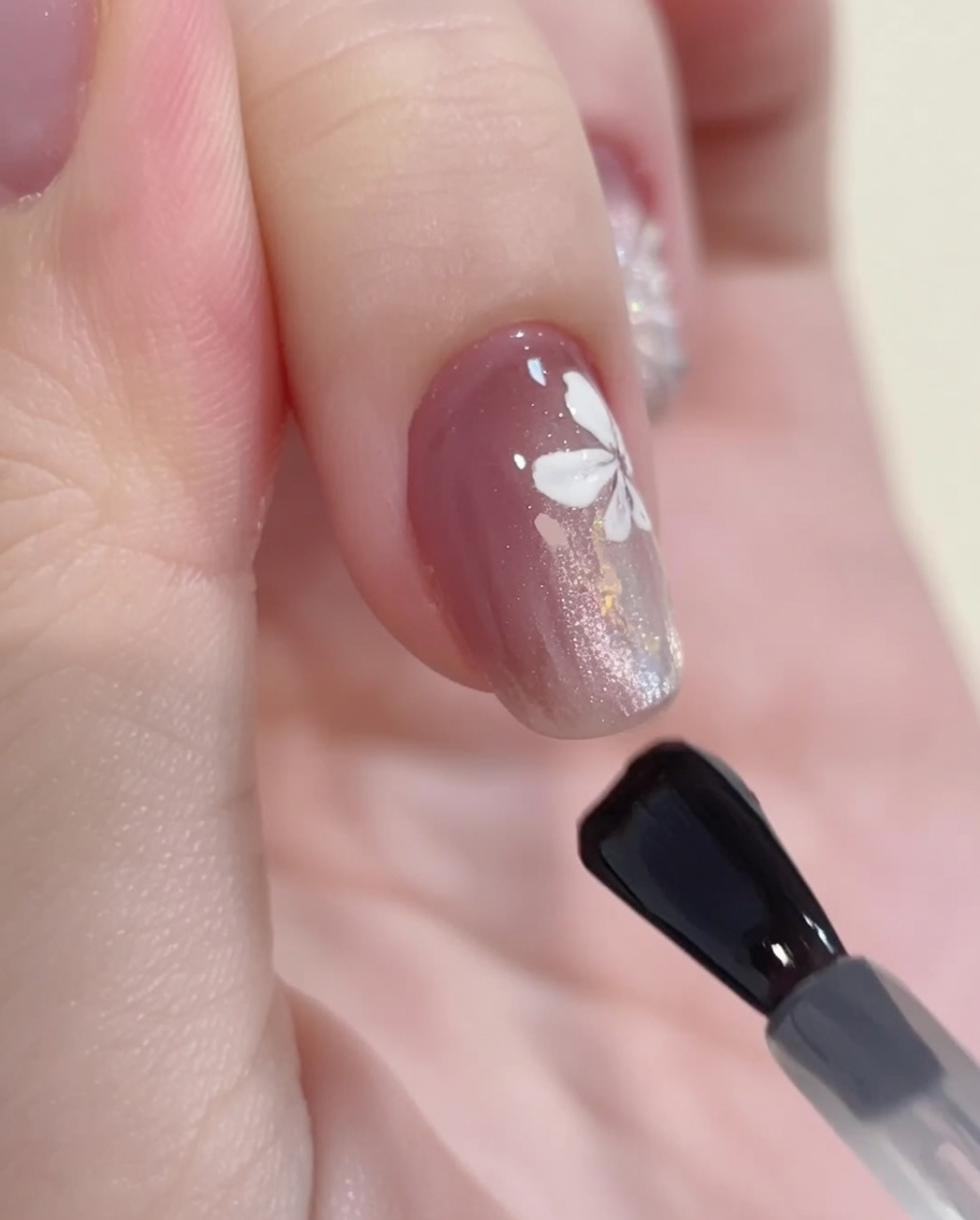 Un pincel de gel aplicando un top coat transparente brillante sobre un diseño finalizado de cat-eye, glitter y arte floral blanco.