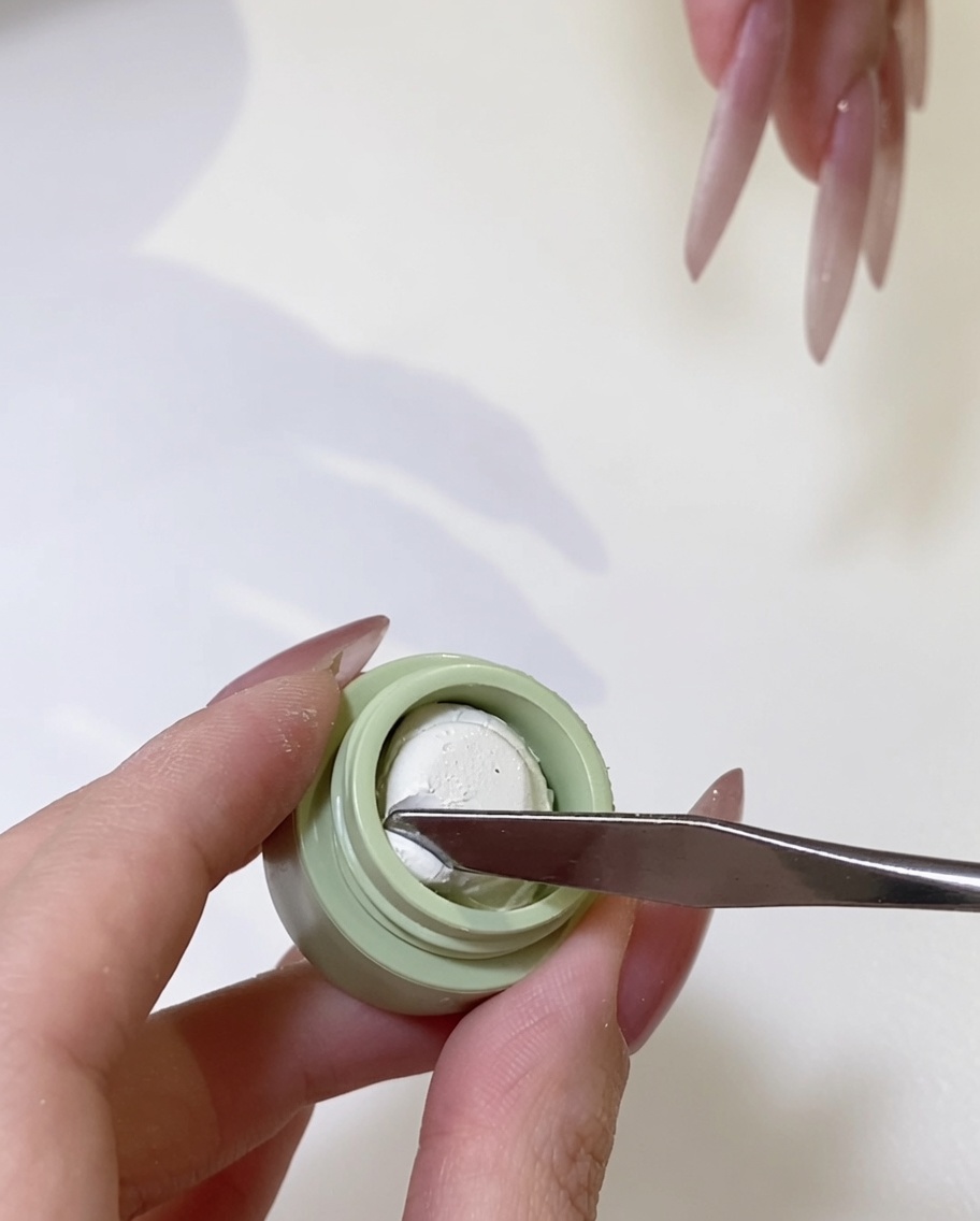Primer plano de una espátula metálica tomando una pequeña cantidad de gel blanco marfil de su contenedor.