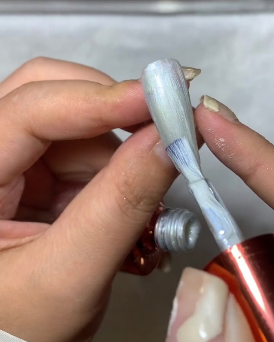 Primer plano de un pincel de uñas aplicando spider gel plateado sobre una uña, creando líneas finas y texturizadas tipo telaraña.