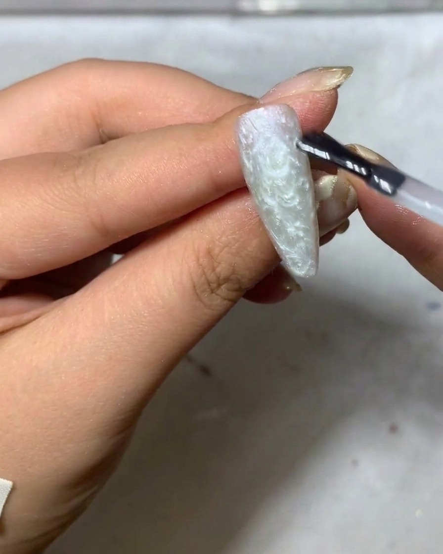Un pincel liner fino comenzando a pintar delicados pétalos de margarita blancos sobre una superficie de uña en tono gris mate.