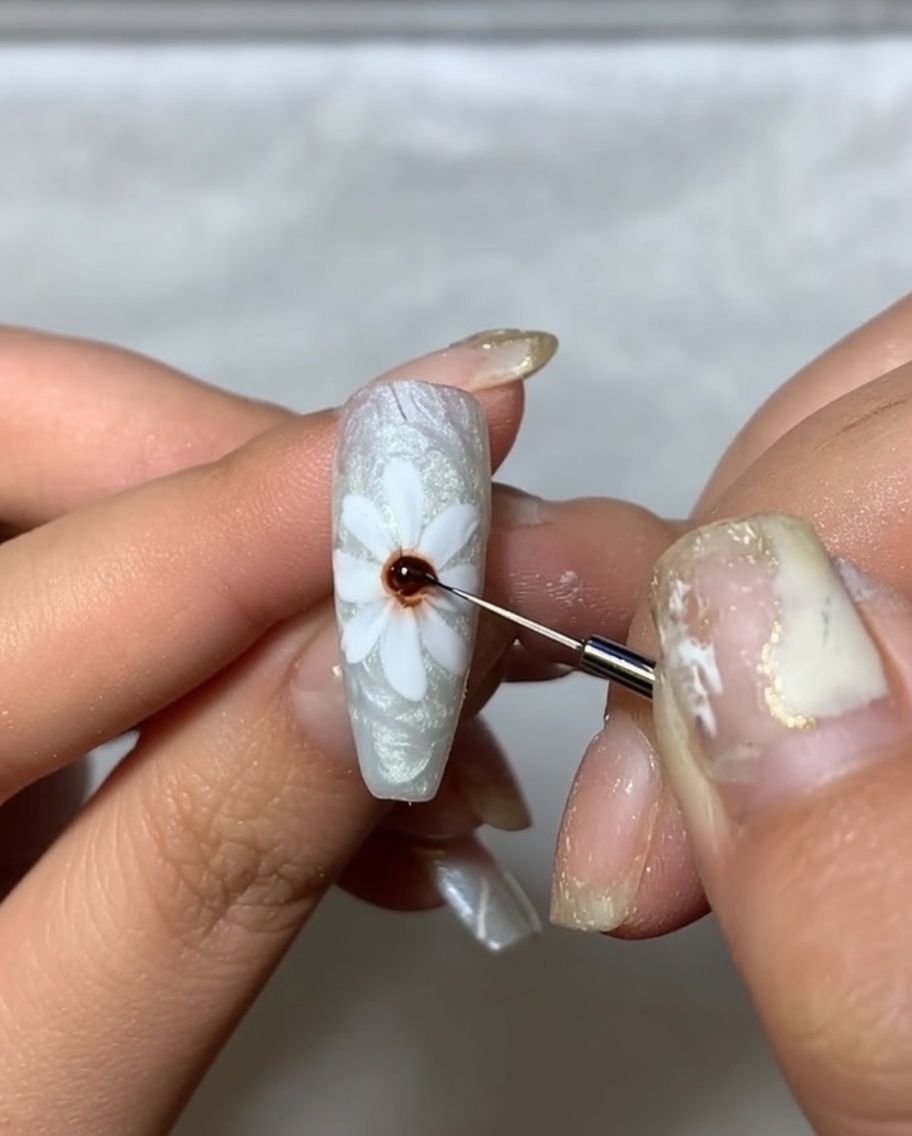 Un primer plano de una uña con pétalos blancos pintados, usando un pincel fino para aplicar un punto de gel marrón oscuro en el centro.