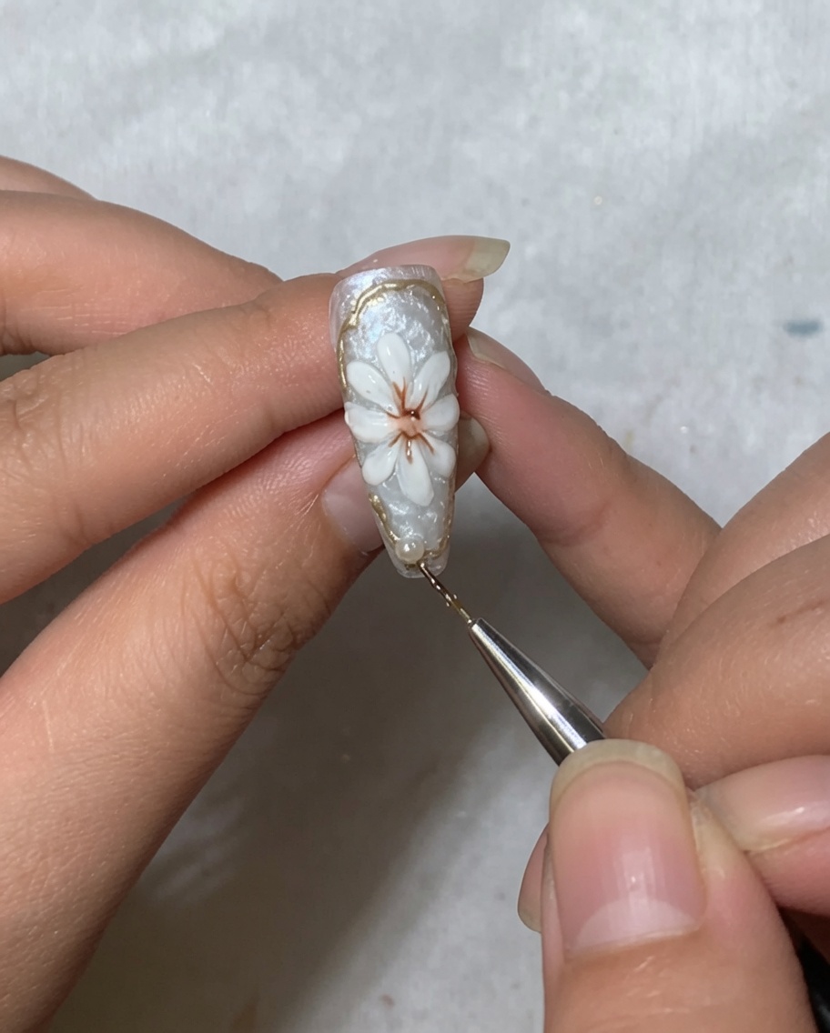 Una herramienta de arte de uñas colocando una pequeña cuenta de perla blanca cerca del área de la cutícula de una uña decorada con una margarita.