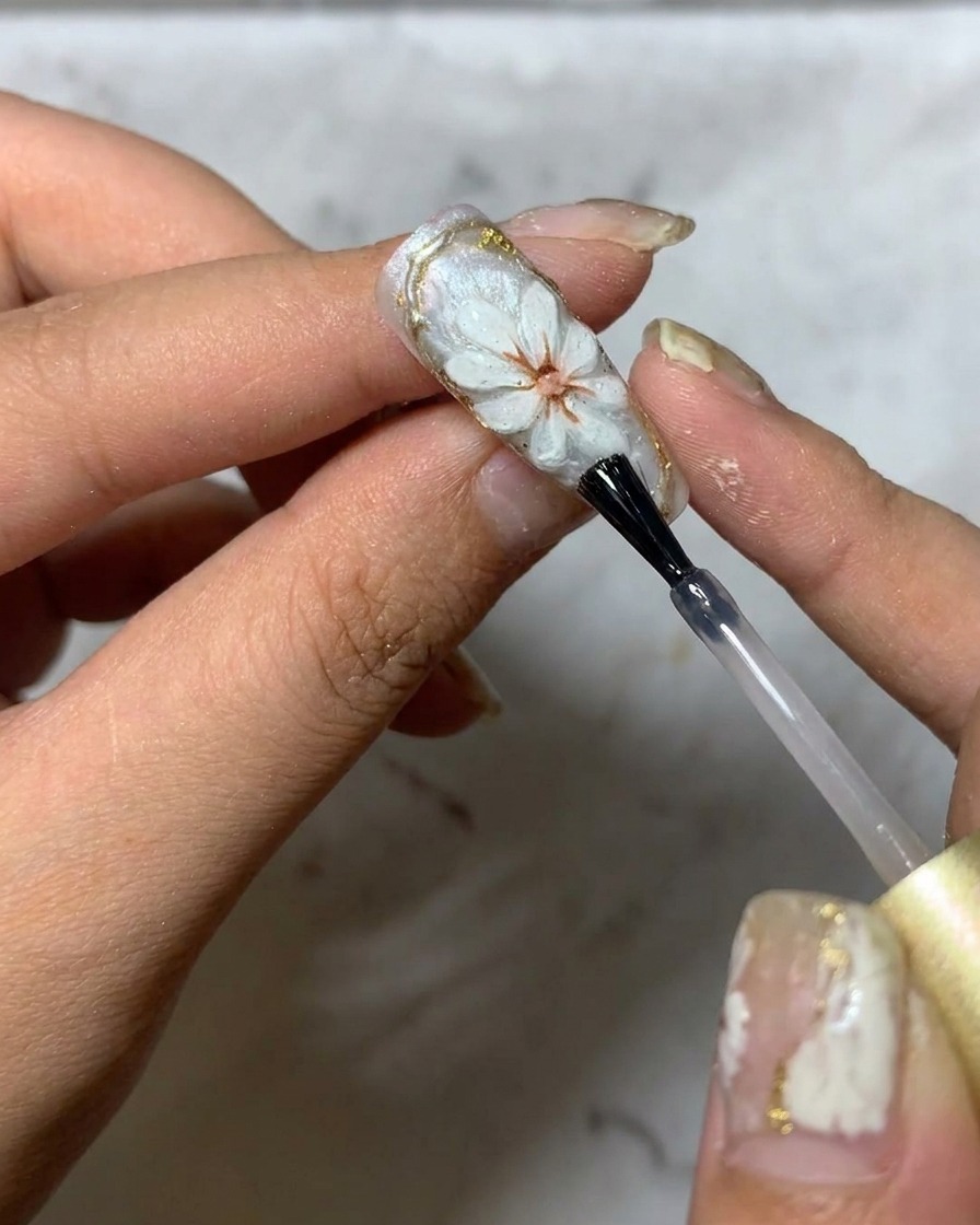 Un pincel aplicando un top coat brillante final sobre un diseño de arte de uñas de margarita en 3D.