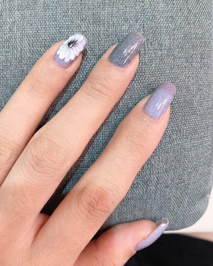 Una mano aplicando una capa de top coat sobre un diseño de uña con una margarita blanca y una pegatina pequeña.
