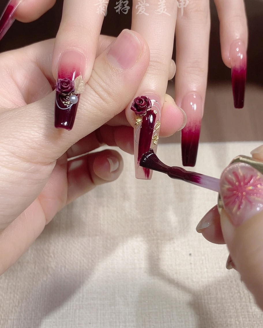 Un pincel fino aplicando un toque de gel con glitter dorado sobre una uña roja.