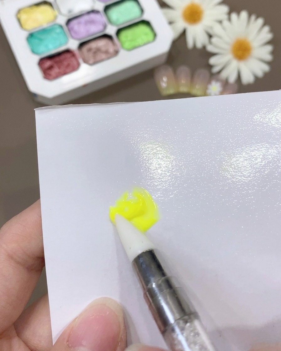 Un pincel mezclando gel amarillo y base coat sobre una superficie blanca para crear una mezcla fina y consistente.