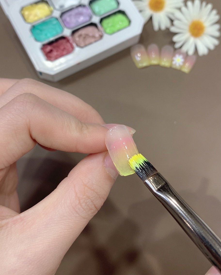 Un pincel aplicando una capa fina de mezcla de gel amarillo transparente sobre la punta de una uña con degradado.