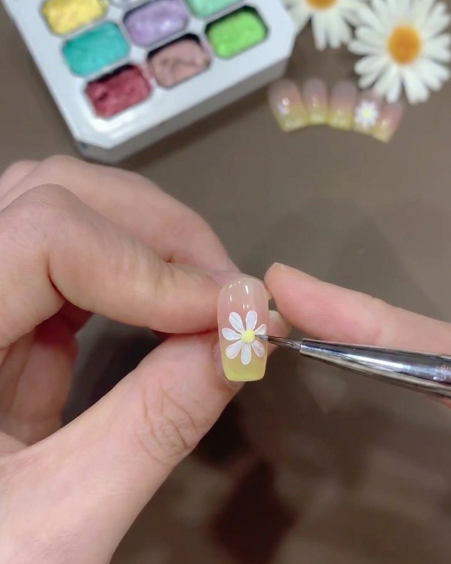 Una pequeña gota de gel amarillo siendo colocada en el centro de una margarita blanca pintada sobre una uña.