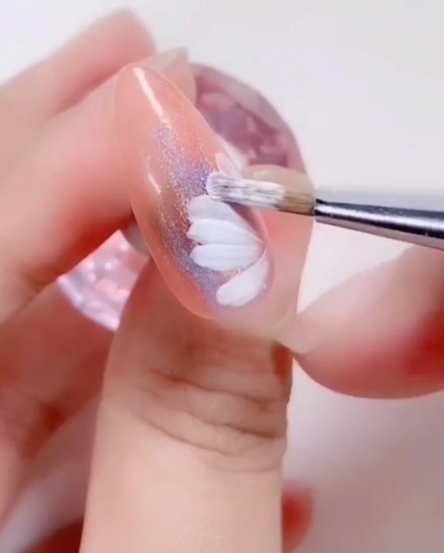 Primer plano de un pincel de uñas pintando pequeños pétalos blancos en el centro interior de un diseño de margarita.