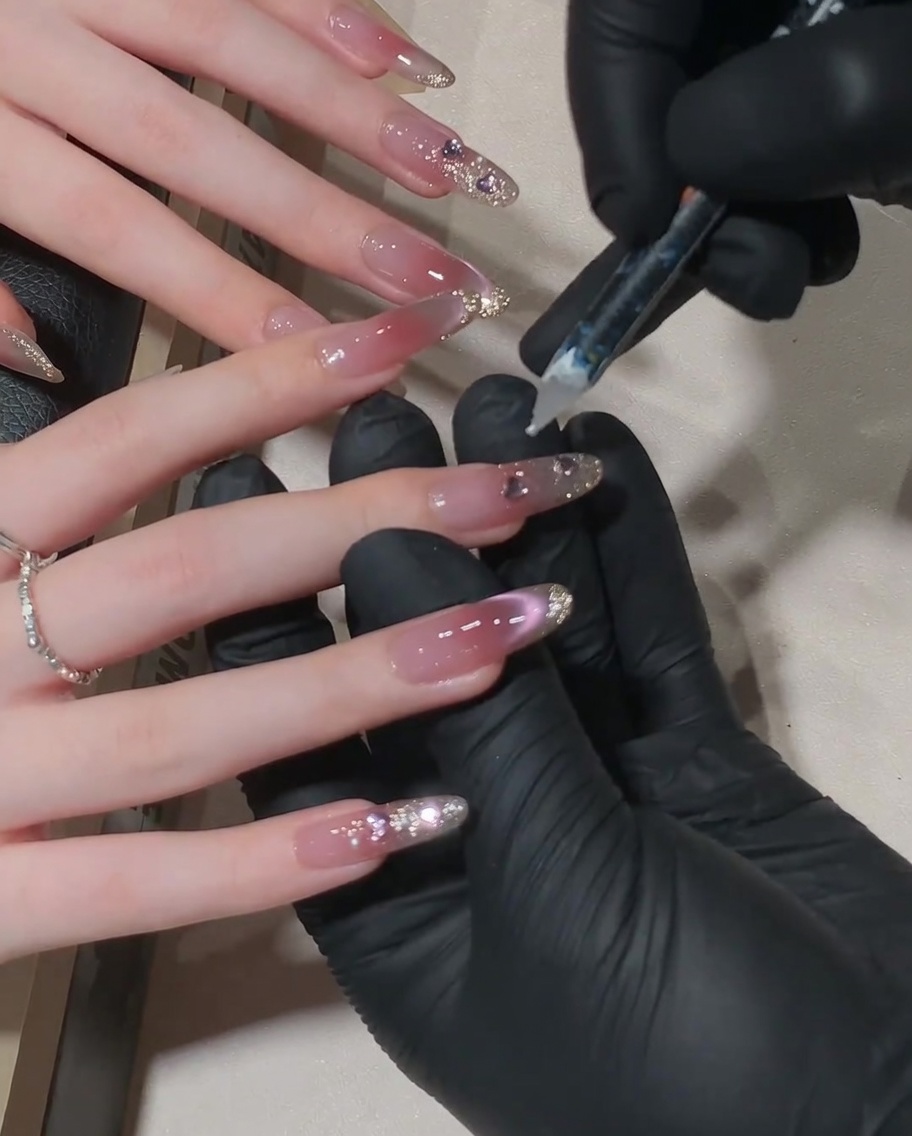Primer plano de un técnico de uñas usando una herramienta con punta de cera para colocar un pequeño cristal en la línea de la cutícula de una uña rosa cat-eye.