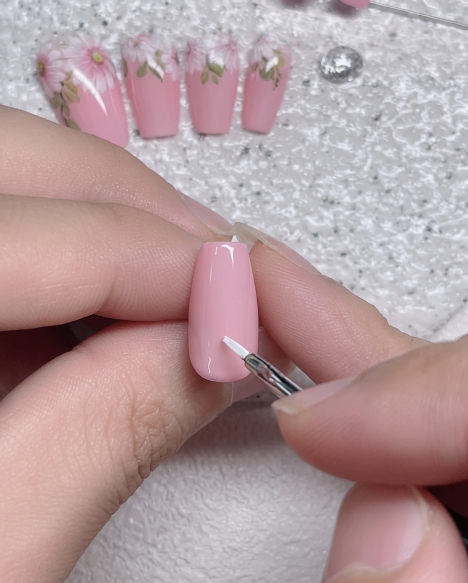 Primer plano de un pincel de detalle fino aplicando un trazo de pétalo blanco sobre una uña con base de gel rosa.