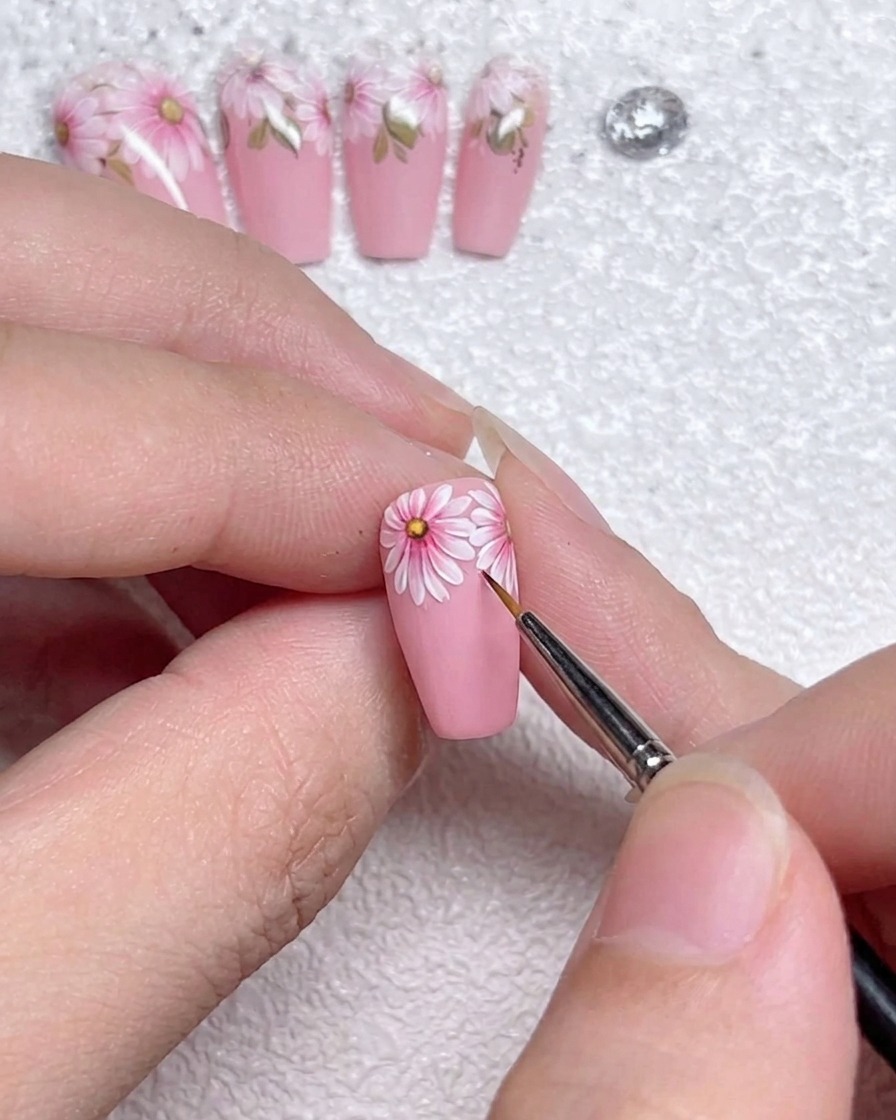 Primer plano de un pincel de detalle fino aplicando líneas de realce blancas precisas en los bordes de los pétalos de margarita sobre una uña pintada con gel rosa.