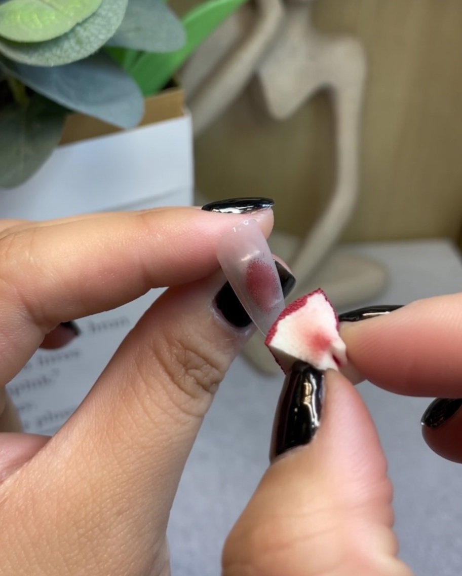 Una esponja de maquillaje aplicando gel rojo vino profundo en el centro de una punta de uña transparente.