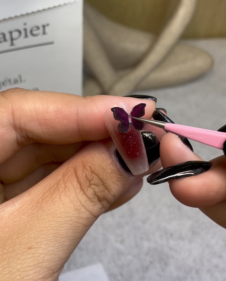 Pinzas colocando un dije decorativo de mariposa burdeos sobre una uña con degradado rojo.