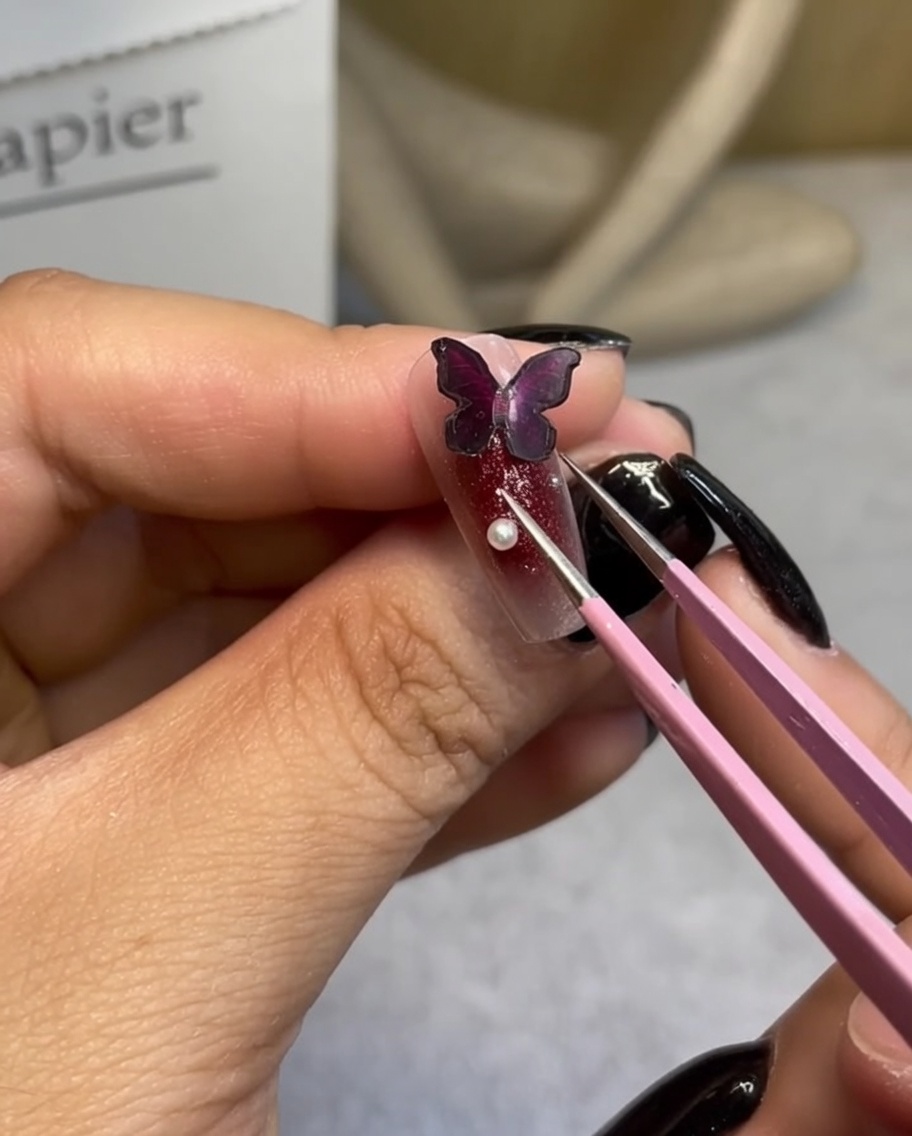 Un par de pinzas de precisión colocando una pequeña perla blanca al lado de un dije de mariposa oscuro en una uña manicurada.