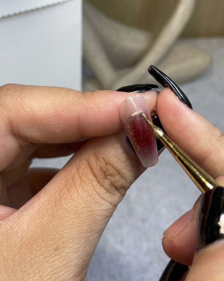 Un pincel de detalle fino aplicando una línea delgada de oro metálico cerca de la cutícula de una uña ombré rojo vino.