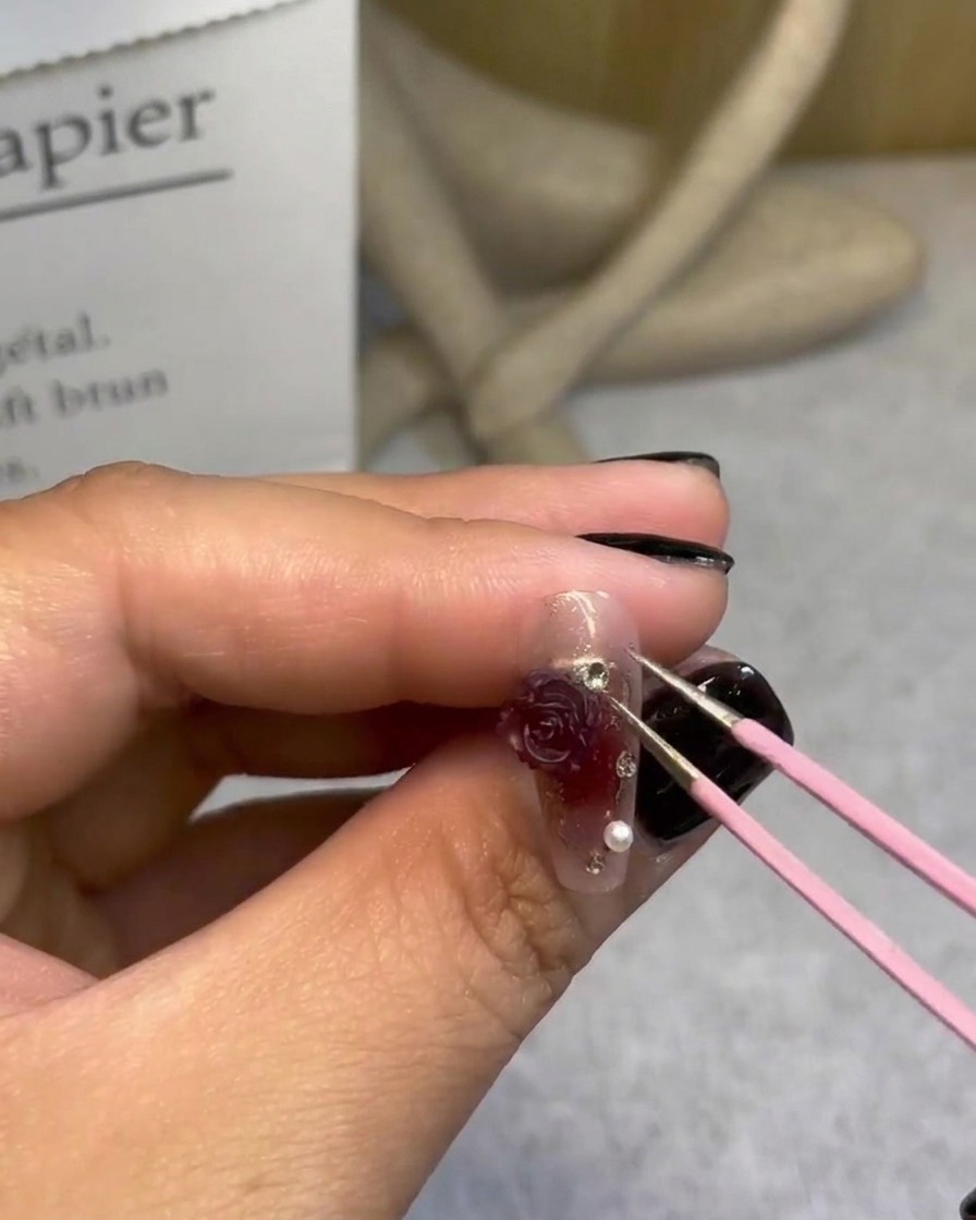 Una mano usando pinzas para agregar pequeñas perlas alrededor de una rosa 3D en un diseño de uña terminado.