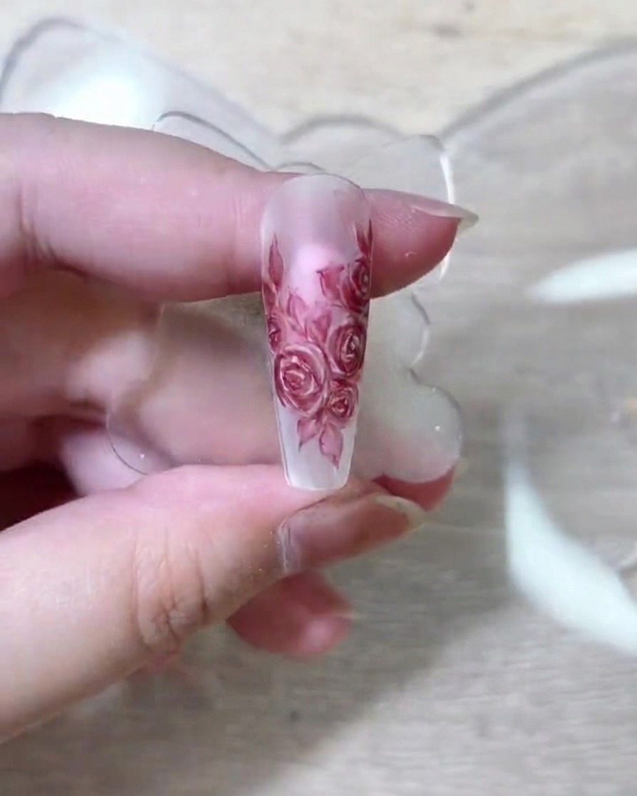 Un pincel de arte detallado depositando pigmento rojo oscuro sobre los pétalos de rosa en una extensión de uña nude.