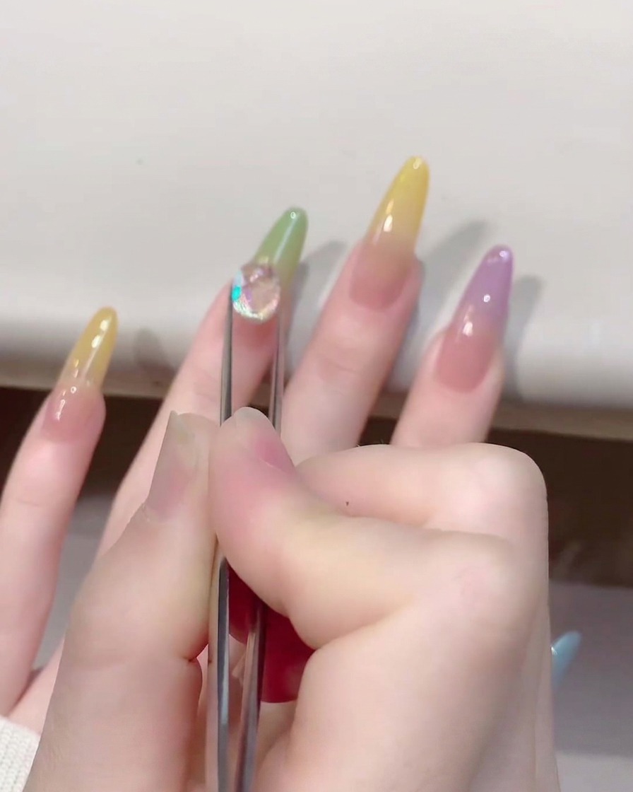 Primer plano de unas pinzas colocando un abalorio brillante y transparente en forma de corazón de cristal sobre una uña color jelly.