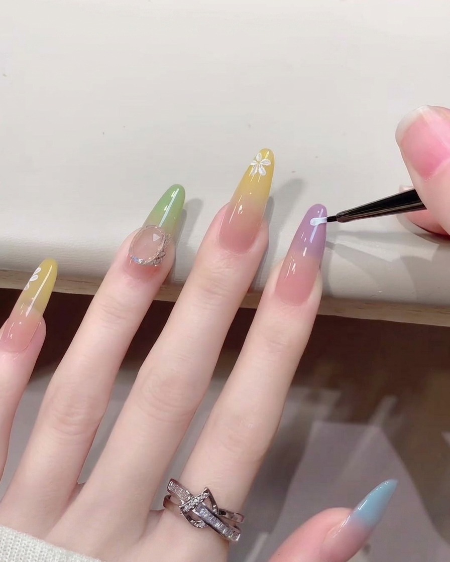 Primer plano de un pincel fino de nail art pintando diminutos pétalos de flores blancas sobre una uña con diseño degradado.