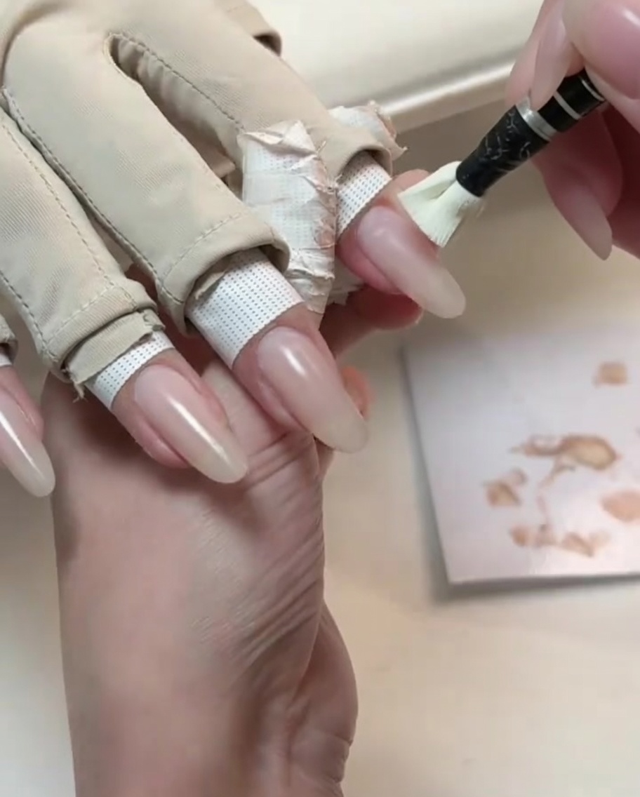 Un pequeño pincel de aplicación cubriendo una uña larga de forma natural con un esmalte en gel nude claro.