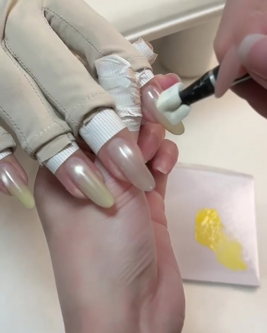 Una herramienta especializada aplicando gel amarillo a toques en la punta de una uña nude con ojo de gato para iniciar un degradado.