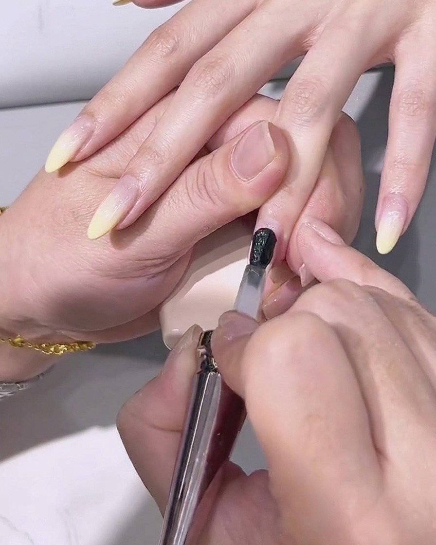 Un pincel aplicando un top coat transparente y brillante sobre la uña con degradado amarillo.