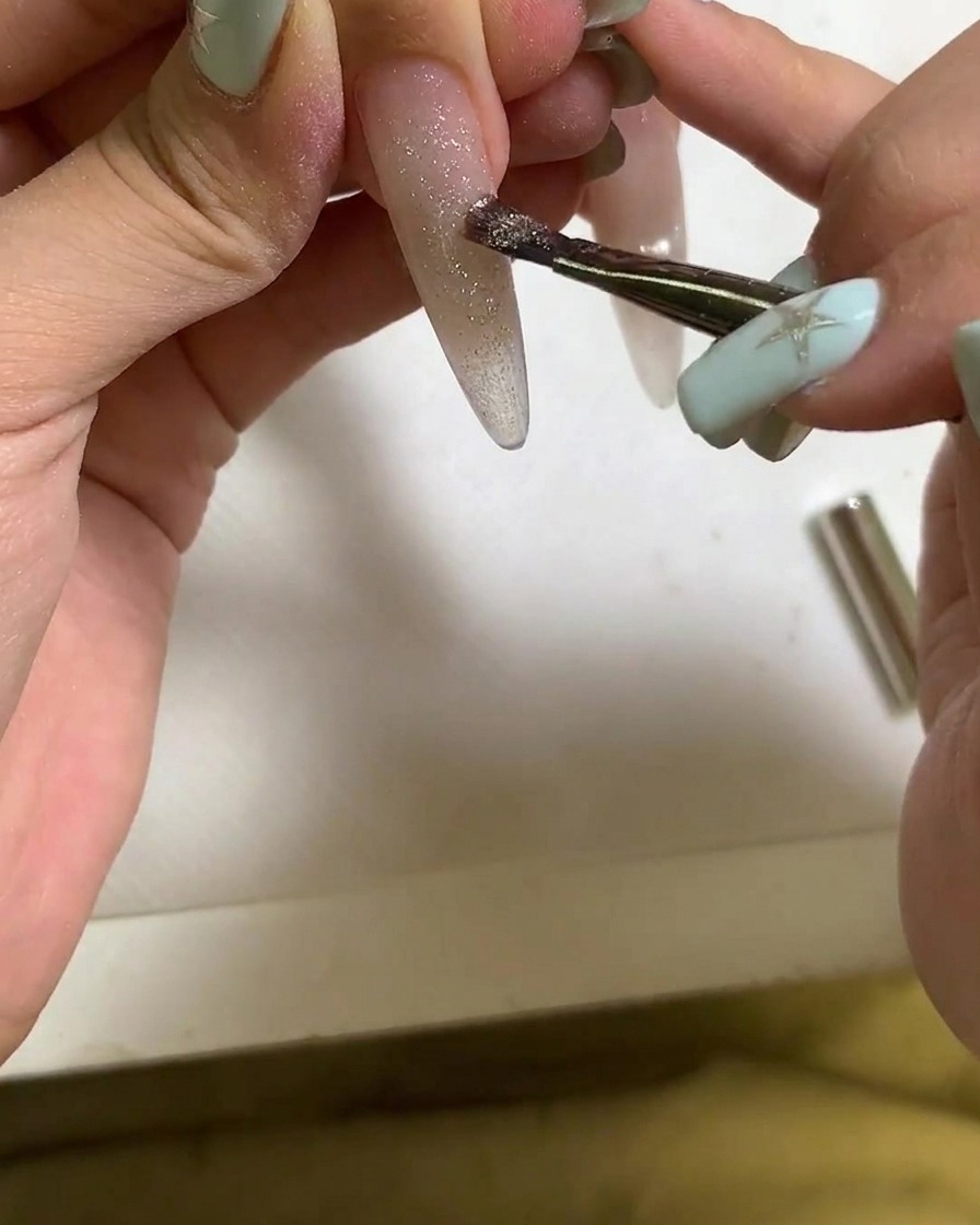 Una brocha aplicando esmalte de gel cat eye sobre una uña alargada.