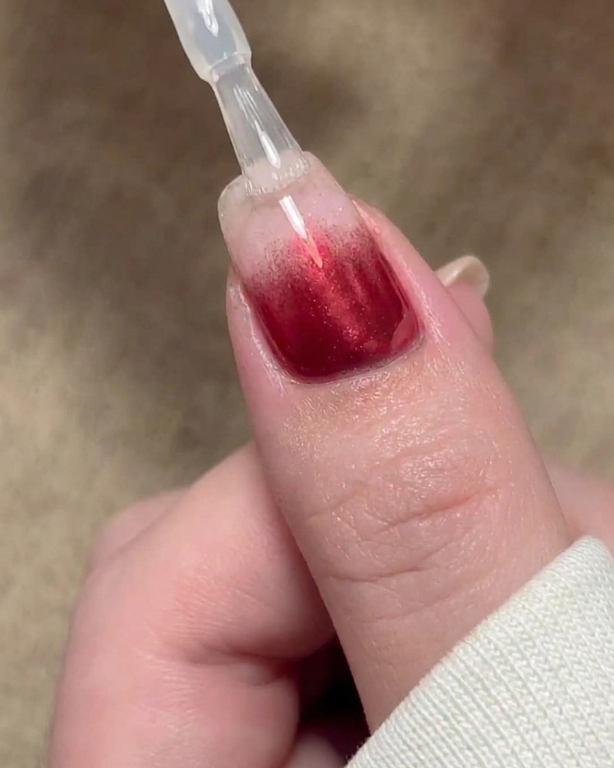Un pincel aplicando una capa selladora de gel base sobre un diseño de uñas con degradado rojo.