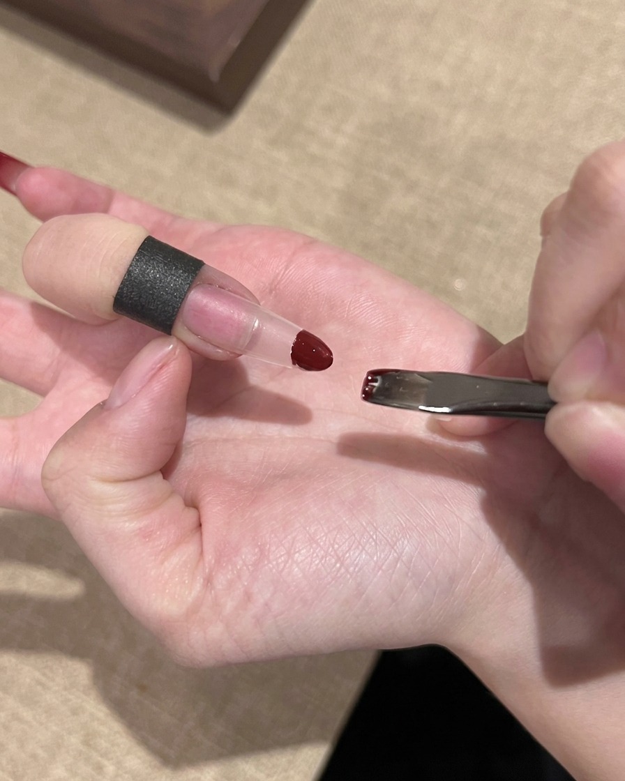 Una mano usando una pequeña esponja para aplicar esmalte de gel rojo en la punta de una extensión de uña transparente.