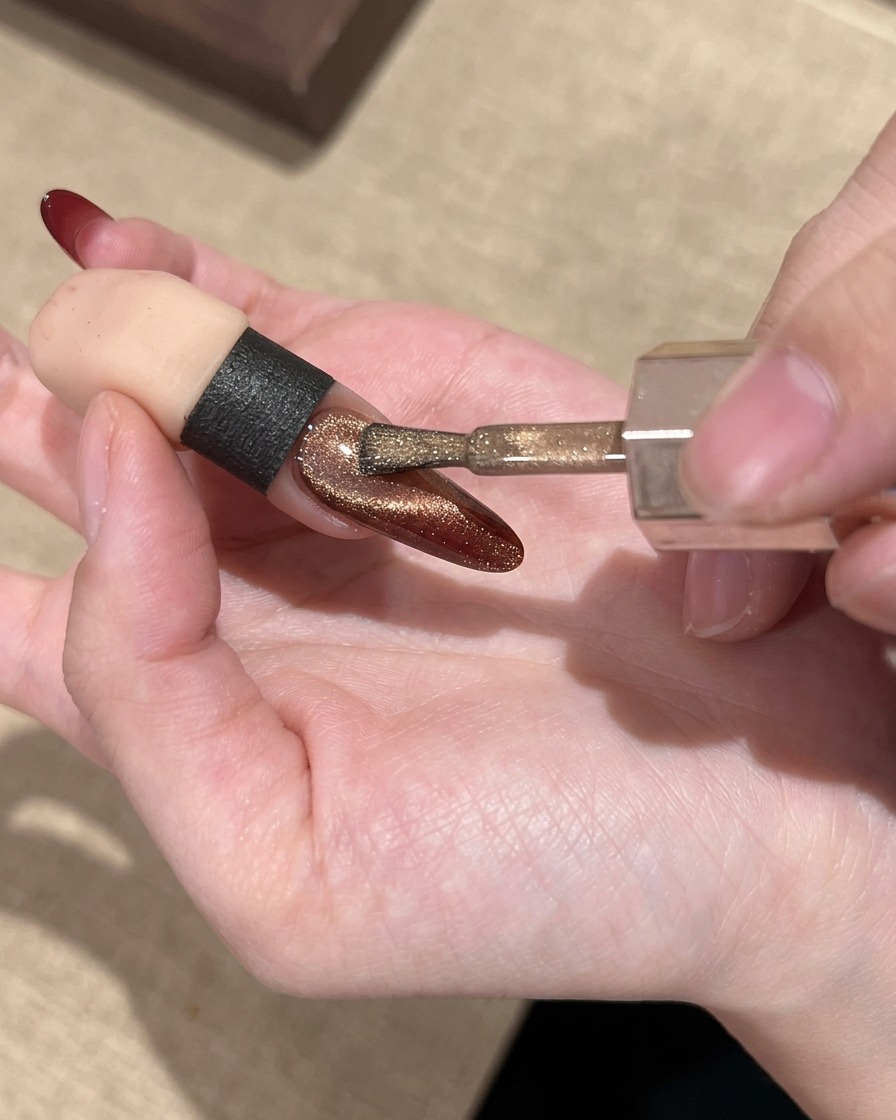 Un pincel de uñas aplicando un gel de ojo de gato dorado brillante sobre la uña con degradado rojo.