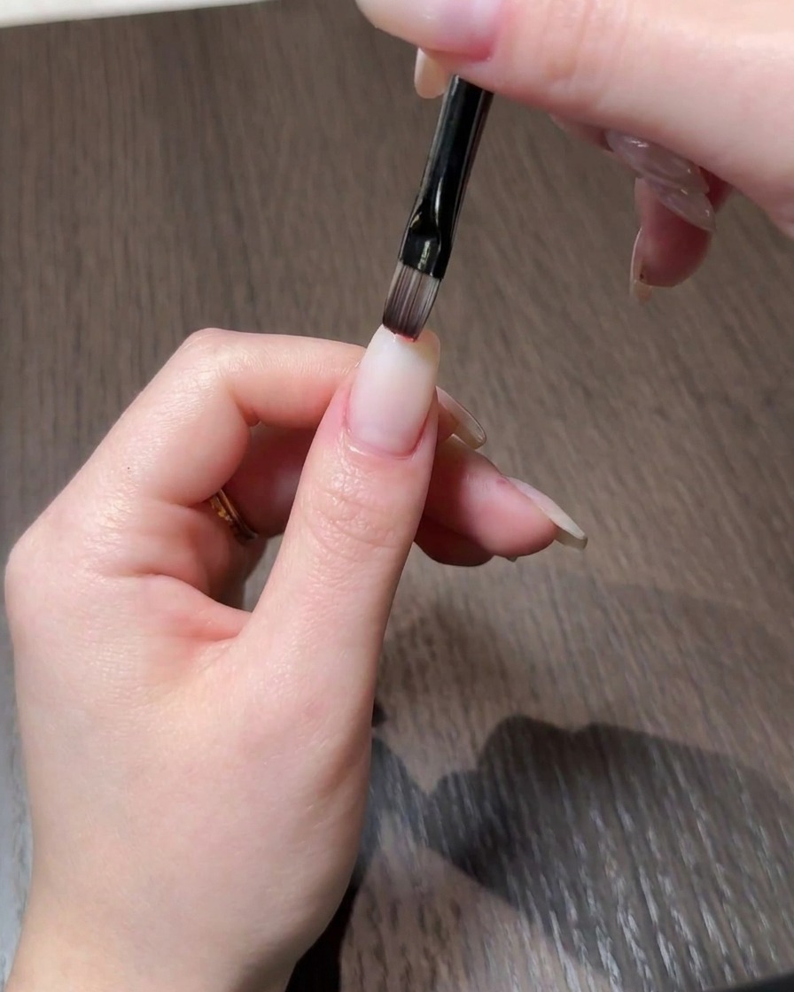Un primer plano de una mano aplicando esmalte en gel rojo en la punta de una uña de aspecto natural usando un pincel fino.