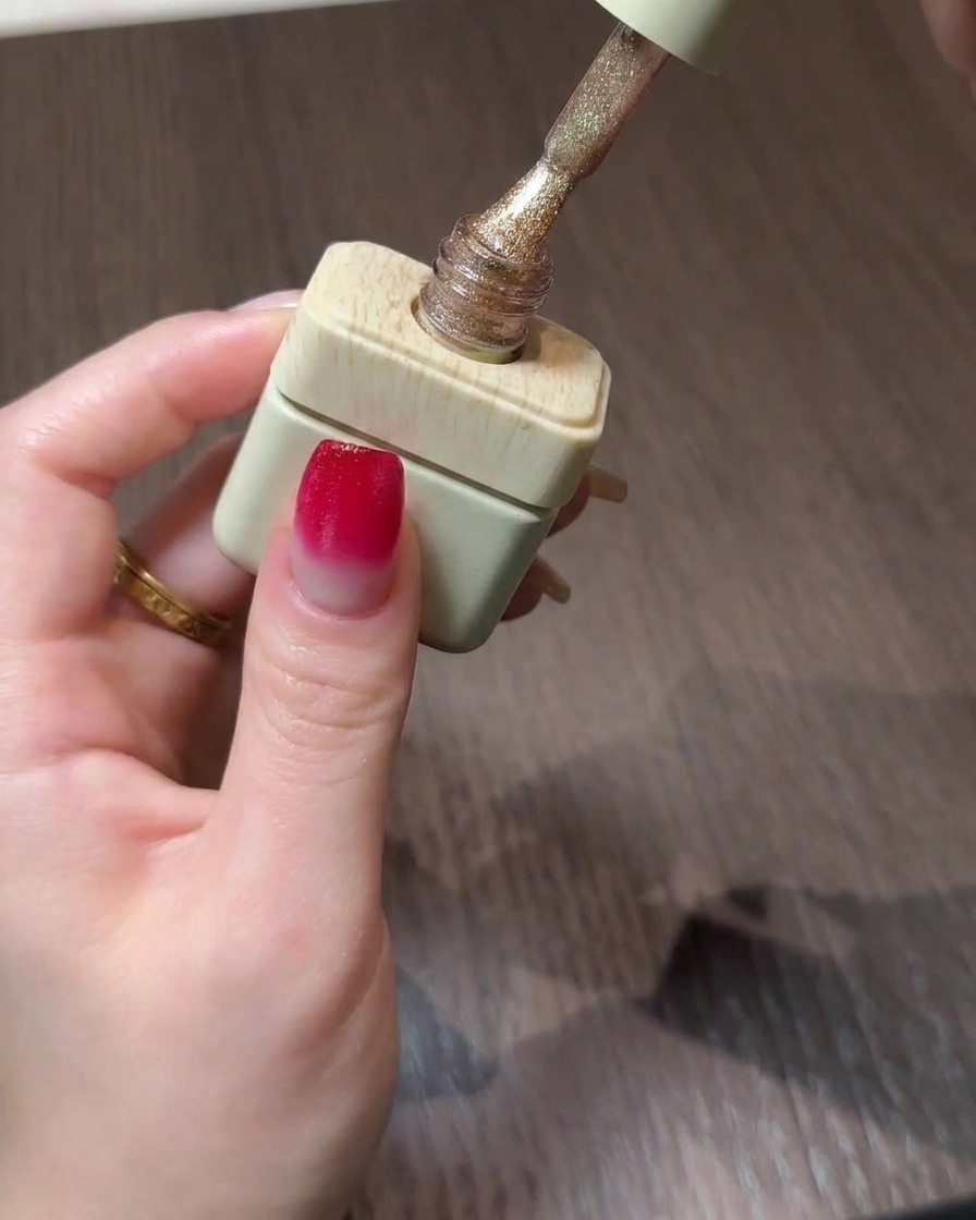 Un frasco de esmalte en gel cat eye dorado con su pincel suspendido sobre una uña degradada.
