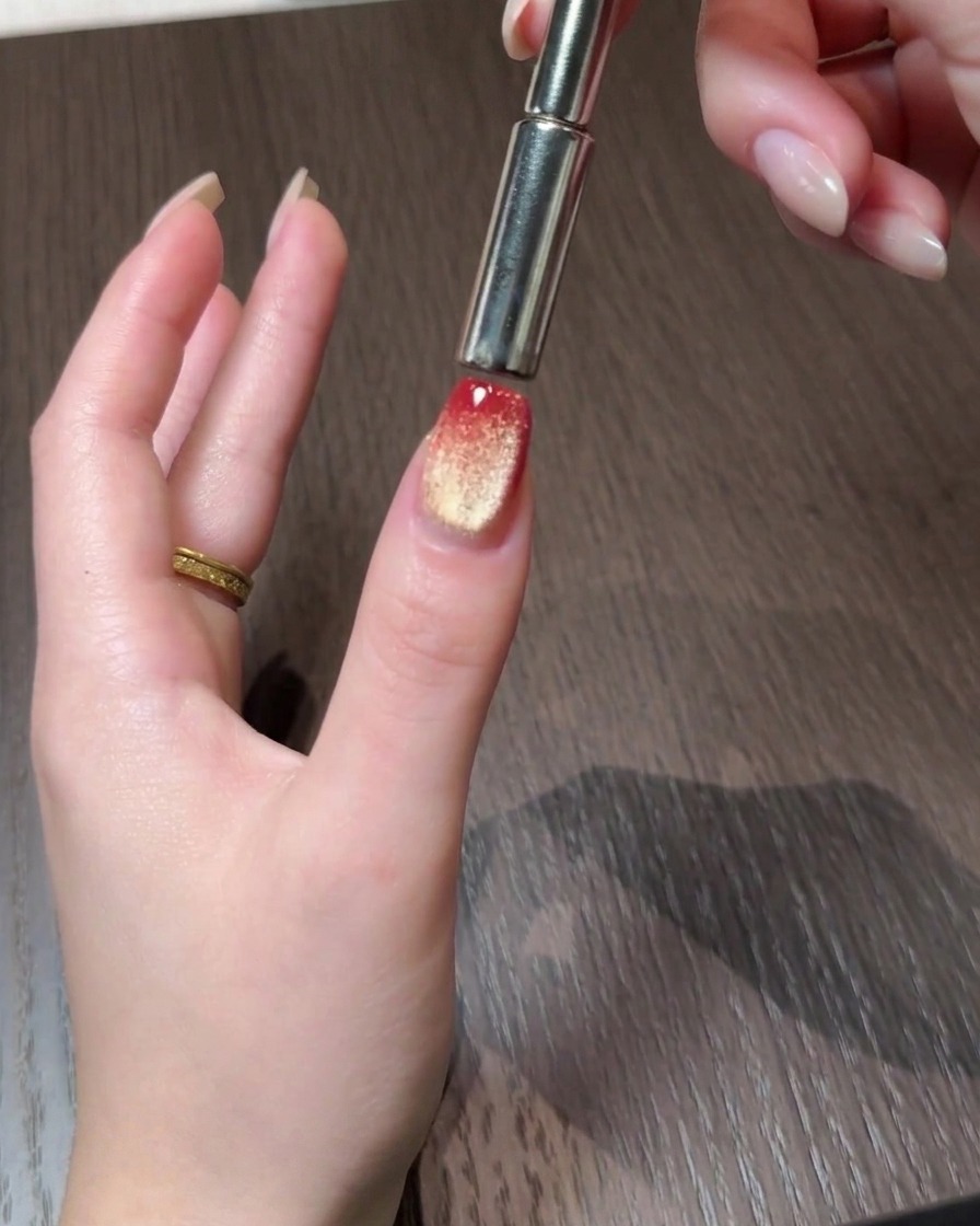 Una herramienta magnética empujando las partículas cat eye hacia el borde posterior de una uña con degradado rojo y dorado.