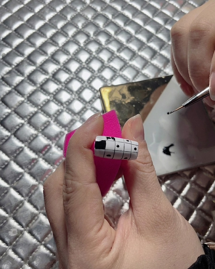 Un primer plano de una artista de uñas usando un pincel delineador fino para enderezar el borde de un cuadrado negro en un diseño de uñas de ajedrez.
