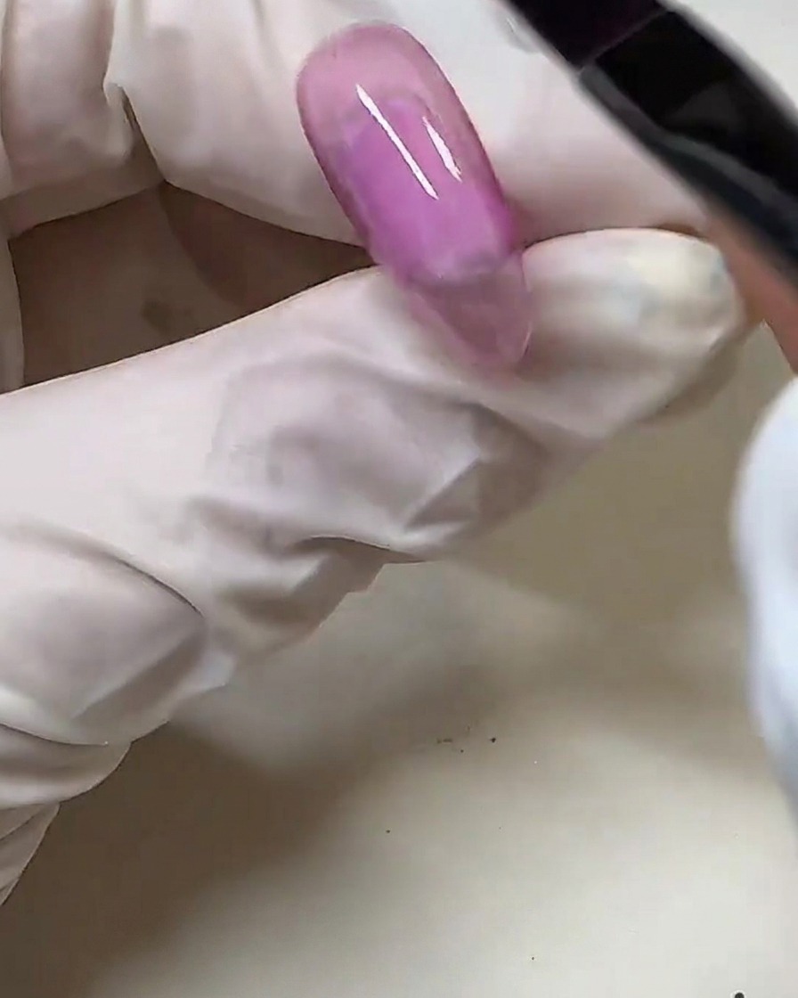 Una mano con guante usando un pincel para aplicar esmalte de gel rosa semitransparente sobre un tip de uña transparente.