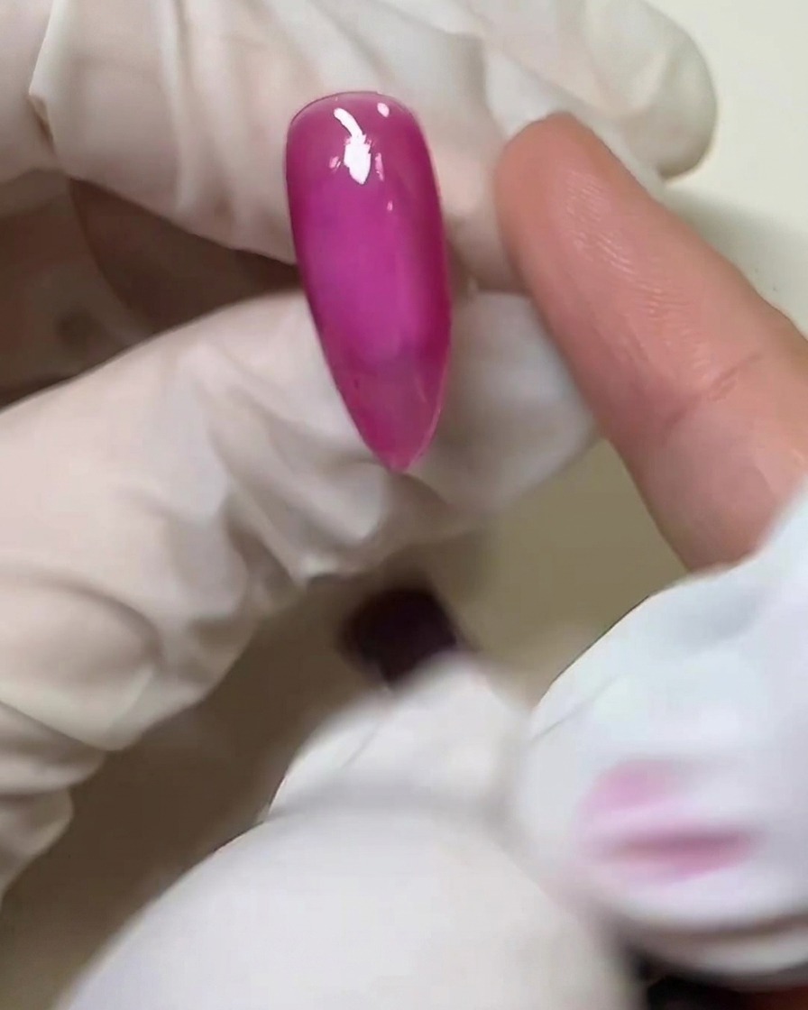 Primer plano de un dedo con guante aplicando una fina capa de pegamento sólido transparente sobre la superficie curada de la uña rosa.
