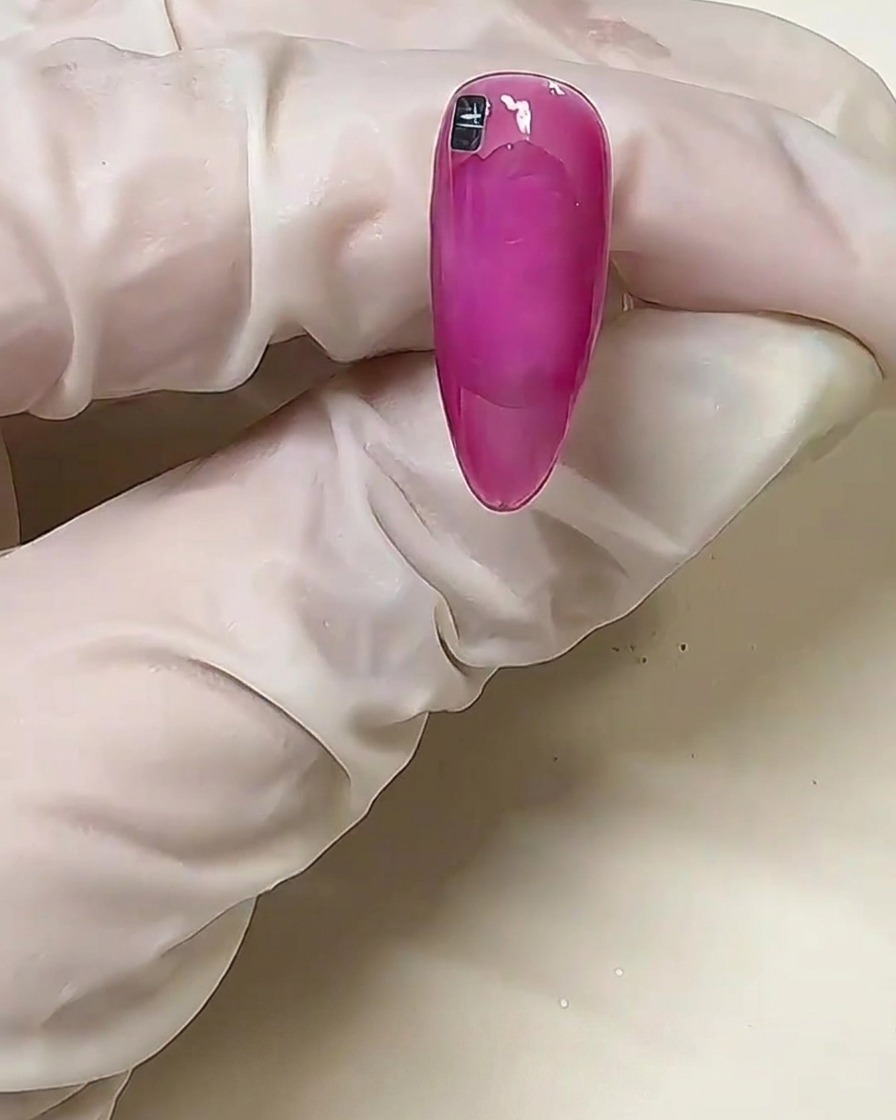 Primer plano de una uña rosa con un solo cuadro metálico plateado colocado cerca de la línea de la cutícula sobre pegamento sólido sin curar.