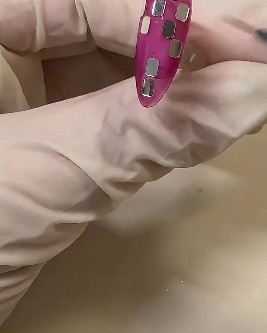 Una mano con guante ajustando pequeños trozos cuadrados de papel plateado en una uña rosa translúcida para crear un patrón de ajedrez.