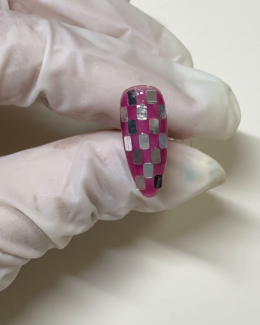 Una mano con guante sosteniendo una uña con un patrón de ajedrez que ha sido cubierta con una capa fina de gel rosa para difuminar los cuadros plateados.