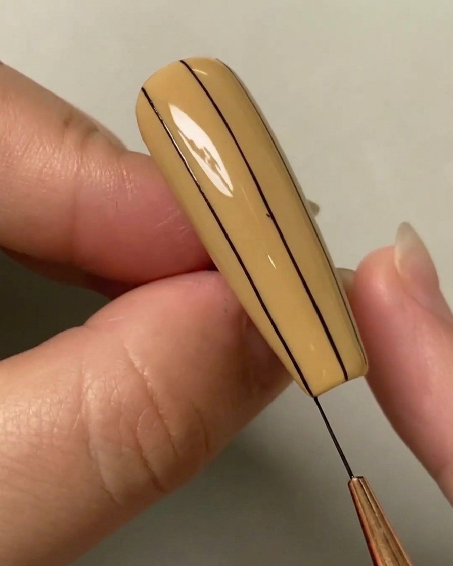 Primer plano de un pincel liner dibujando una tercera línea vertical negra en el lado derecho de una uña color nude.